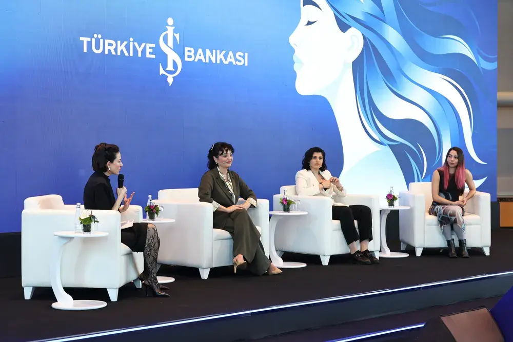 İş Bankası logosunun ve mavi saçlı stilize bir kadın profilinin yer aldığı büyük mavi bir dijital ekranın önündeki sahnede, beyaz koltuklarda oturan dört kadın konuşmacı, mikrofonla konuşan birinin önderliğinde Sınırların Ötesinde bir panel tartışması gerçekleştiriyor.